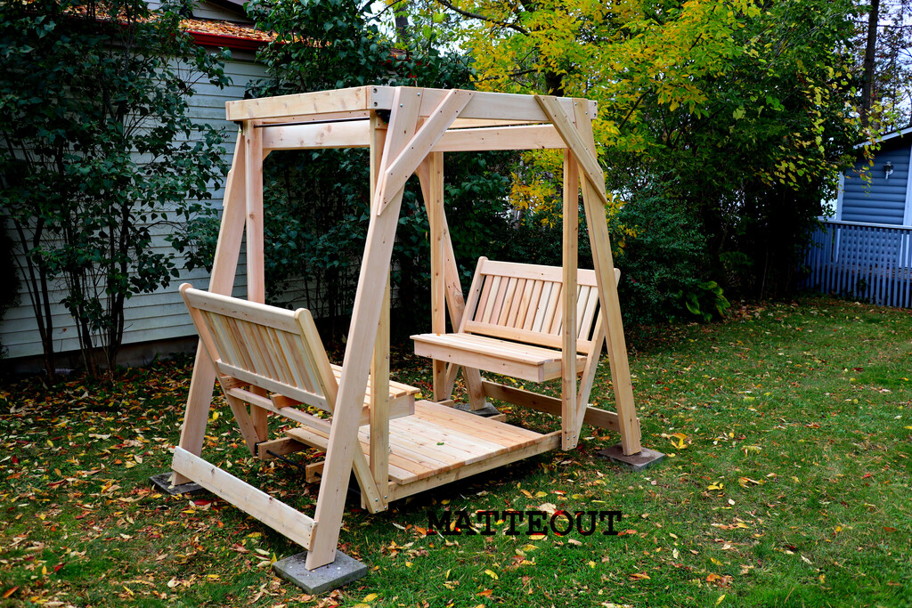 Matteout handcrafted 4-person wooden glider swing with an overhead structural frame set up on concrete blocks in a backyard lawn.