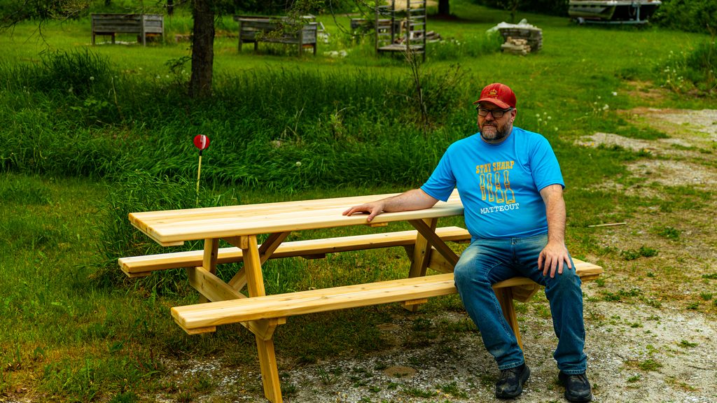 Local artisan builder sitting on a handcrafted heavy-duty wooden rectangular picnic table wearing a Matteout brand t-shirt.